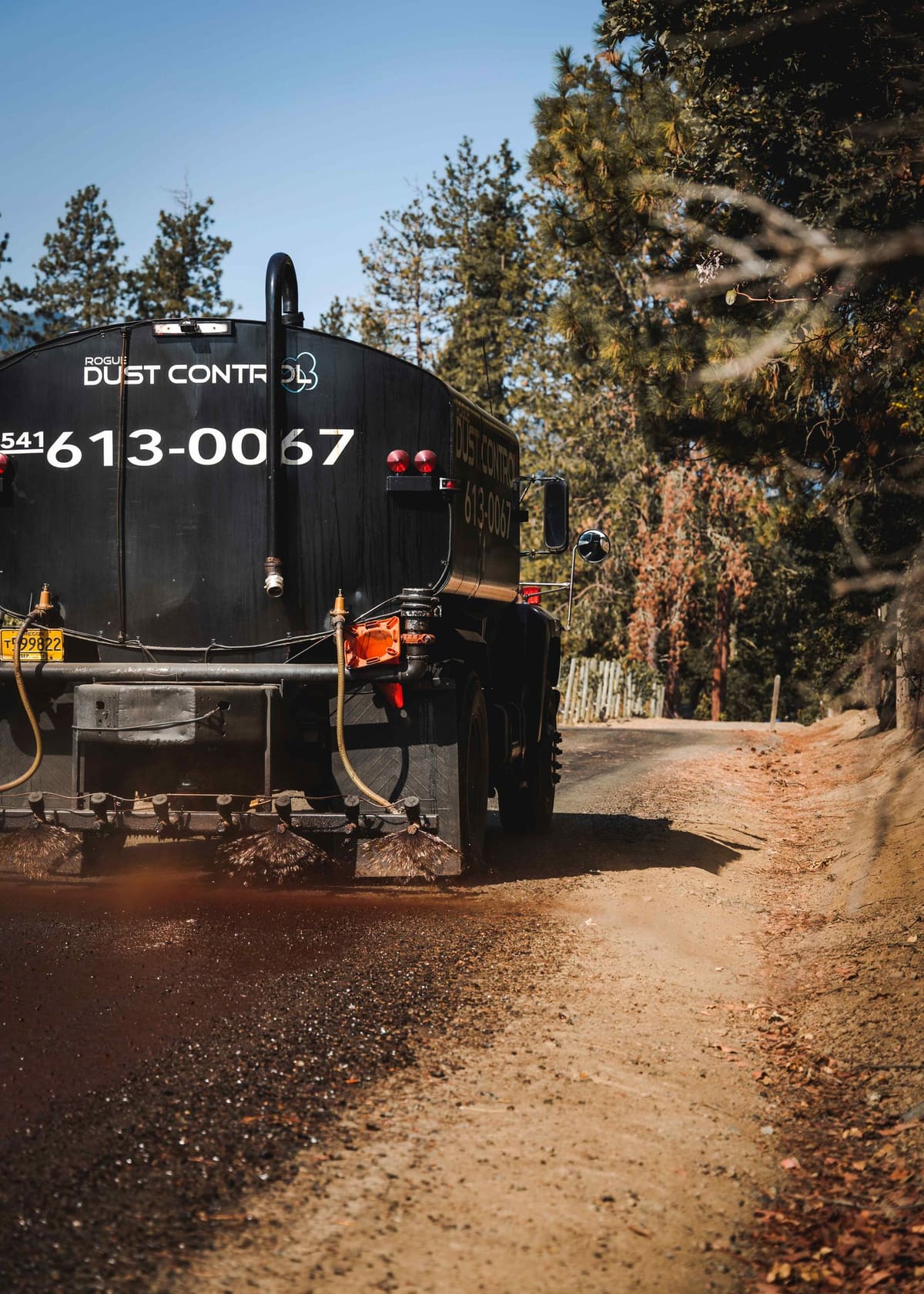 The black tanker driving away down a dirt road through pines, spraying as it goes.