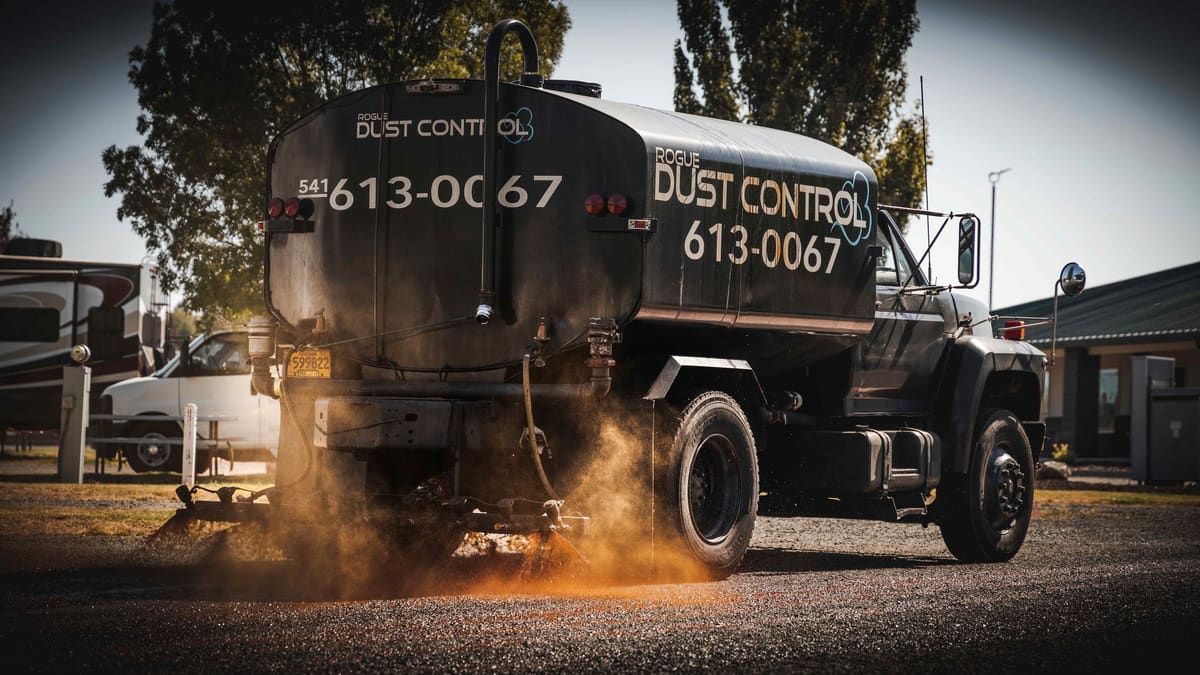 Rogue Dust Control's black Ford tanker spraying a gravel lot in late-afternoon sun, 613-0067 lettered on the tank.