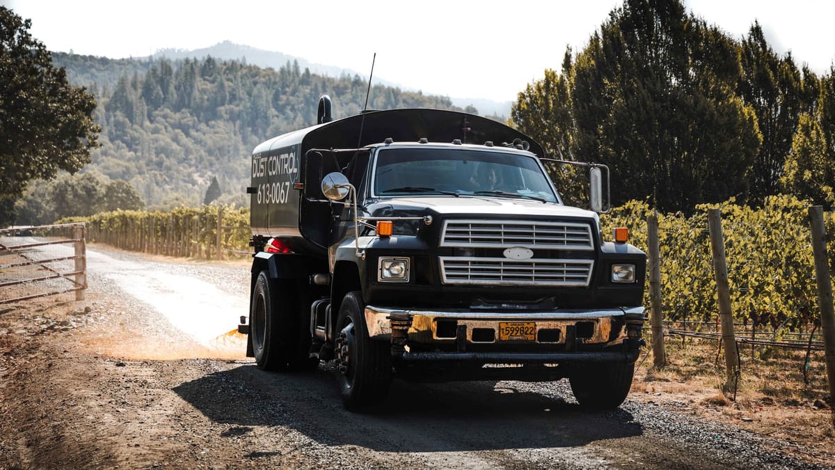 Black Rogue Dust Control truck on a treated gravel road between vineyard rows.