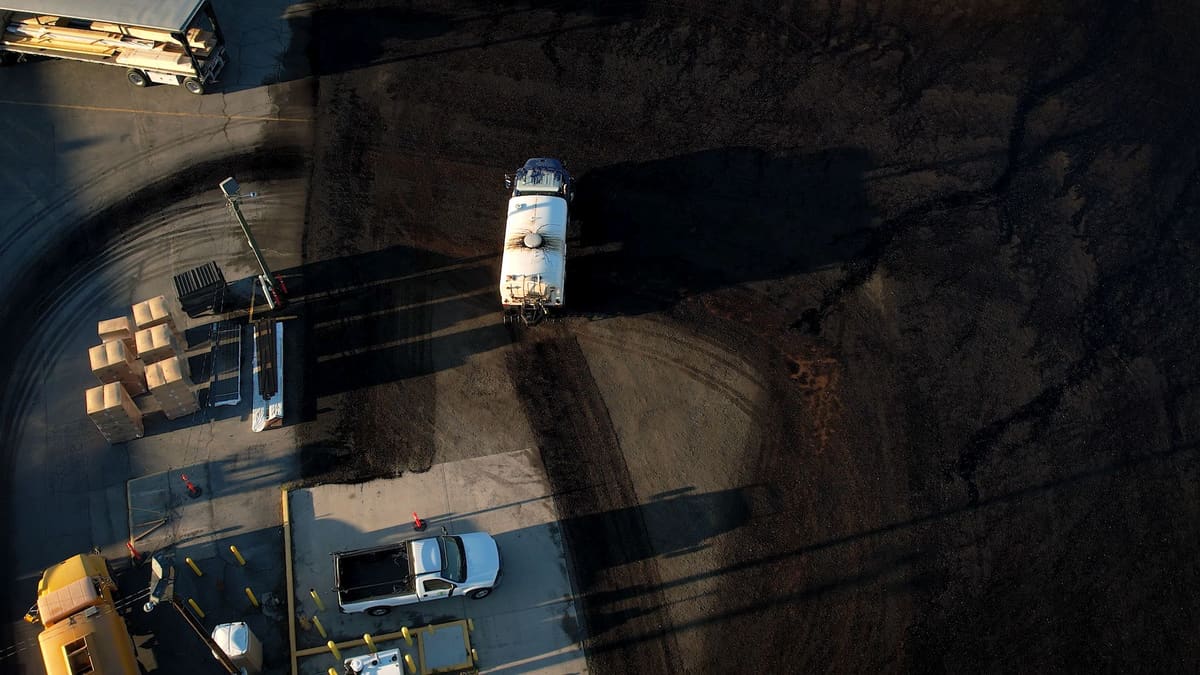Straight down from the drone: a Rogue Dust Control tanker crossing a dark, freshly treated yard, a pickup and stacked materials at the edge of the lot.