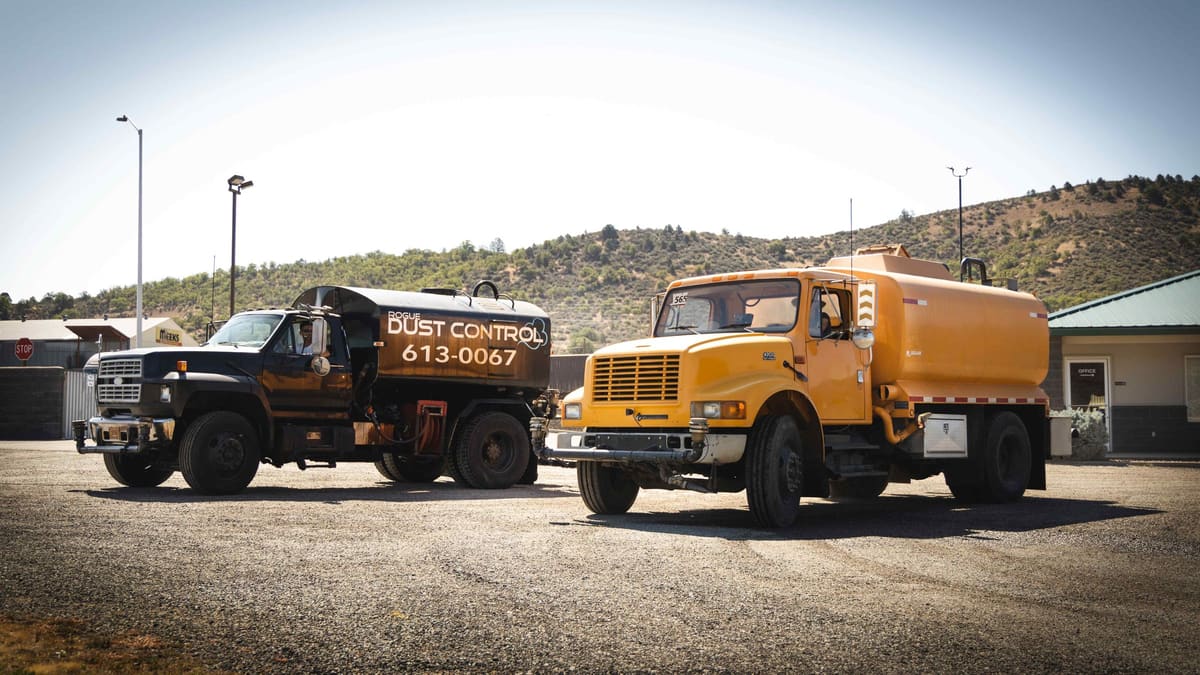 Two of Rogue Dust Control's trucks, the black tanker and the yellow tanker, parked side by side in a gravel lot.