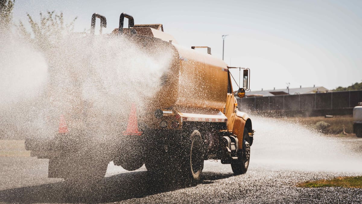 The yellow tanker spraying a gravel surface, water fanning out from the spray bar.